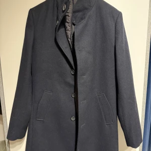 Mörkblå rock från Bruun & Stengade - Snygg mörkblå rock från Bruun & Stengade med klassisk rak passform och knappar framtill. Jackan har två fickor på sidorna och quiltad, grå innerdel som ger extra värme. Perfekt för dig som gillar stilrena och tidlösa plagg.