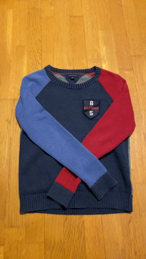 Blå och röd stickad tröja Tommy Hilfiger - Snygg stickad tröja från Tommy Hilfiger i marinblått, rött och blått. Perfekt för dig som gillar sportig stil. Den är även bra för att ha skjorta under.
