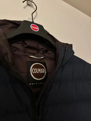 Mörkblå Colmar pufferjacka med huva - Tjena säljer nu min otroligt snygga Colmar jacka då den inte kommer till användning, den är i mycket bra skick och är ej använd mycket, den har inga fysiska hål eller slitningar, köpte den förra vintern men den har inte använts mycket alls så därför jag säljer. Jag säljer då en mörkblå pufferjacka från Colmar med huva och svarta dragkedjor. Jackan har två sidofickor med dragkedja och ett brunt innerfoder. Perfekt för dig som vill ha en stilren och varm jacka med snygg passform.