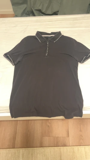 Svart piké från Armani Exchange - Svart pikétröja från Armani Exchange med vit logga längs dragkedjan och vita detaljer på krage och ärmslut. Klassisk passform och kortärmad, perfekt för en clean och stilren look.