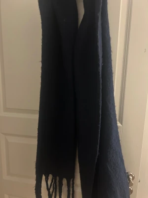 Mörkblå ullhalsduk med fransar - En stilren mörkblå halsduk i ull med klassiska fransar i ändarna. Perfekt att svepa runt halsen för extra värme under kyliga dagar. 