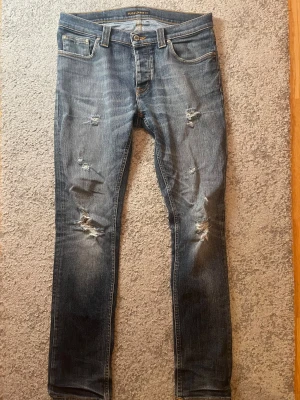 Blå slitna jeans från Nudie Jeans - Säljer ett par blå jeans från Nudie Jeans med snygga slitningar och hål på benen för en cool, avslappnad look. Klassisk femficksmodell med orange sömmar och Nudie-logga på bakfickan. Jeansen har rak passform och är tillverkade i bomull.