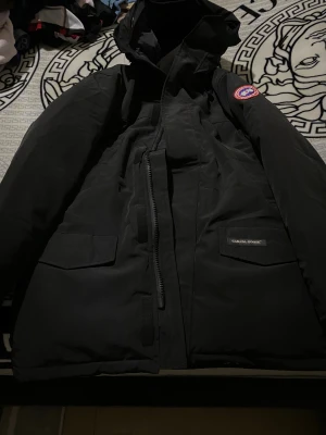Svart dunjacka Canada Goose XL - Svart dunjacka från Canada Goose i storlek XL. Jackan har huva, två stora fickor med lock, dragkedja framtill och klassisk Canada Goose patch på ärmen. Materialet är slitstark polyester och jackan är perfekt för kalla vinterdagar.