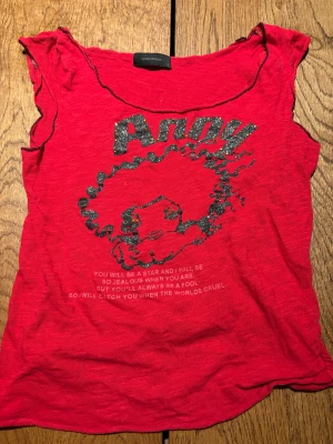 Y2K Röd topp med glittertryck från Vero Moda - Cool röd topp från Vero Moda i storlek S med glittrigt tryck och text framtill. Motivet har silvriga glitterdetaljer och små guldiga paljetter. Toppen har korta ärmar med lätt rynk och är gjord i mjuk bomull. Perfekt för dig som gillar statement-plagg!