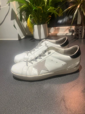 Golden Goose Superstar vita sneakers - Golden Goose Superstar sneakers i vitt skinn med grå mockadetaljer och snörning. Ikonisk stjärna i grått på sidan och svart hälparti med logotyp. Rund tå och platt sula ger en klassisk och cool look. Perfekt för dig som gillar streetstyle och exklusiva märken. Sulan är i förvånansvärt bra skick för o vara Golden goose  inte sönder utan helt intakta. Kontakta vid funderingar