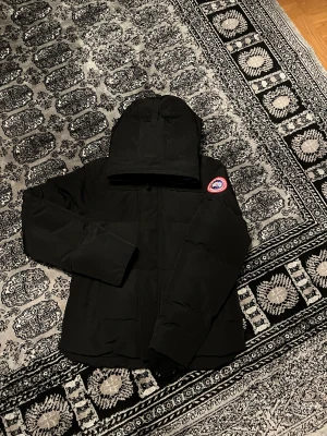 Svart dunjacka från Canada Goose - Svart dunjacka från Canada Goose med huva och klassisk logotyp på ärmen. Jackan har dragkedja framtill, snörning vid huvan och quiltad design. Perfekt för kalla dagar och har en clean, stilren look som funkar till allt. Flera fickor och snygga detaljer.