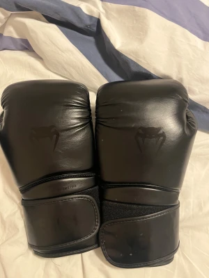 Svarta boxningshandskar från Venum - Säljer ett par svarta boxningshandskar från Venum med coolt logotryck på ovansidan. Handskarna har en rundad form, bred kardborreknäppning och är tillverkade i syntetmaterial med slät finish. Perfekta för träning och sparring.