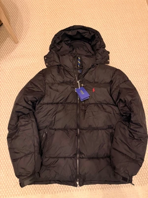 Svart pufferjacka från Polo Ralph Lauren - Svart pufferjacka från Polo Ralph Lauren med huva, RL-dragkedja och röd broderad logga på bröstet. Jackan har quiltad design och två sidofickor med dragkedja. Perfekt för kalla dagar och har en clean, sportig look.