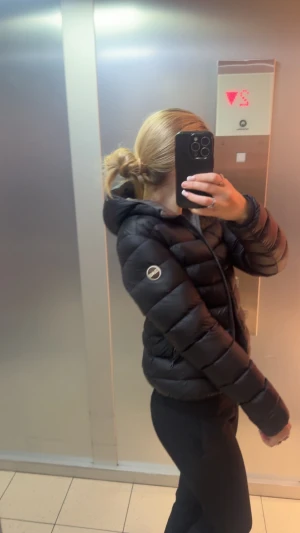 Svart pufferjacka från colmar  - Säljer en svart pufferjacka från colmar med huva och dragkedja framtill. Jackan har ett stilrent, quiltat mönster och logotyp på ärmen. Perfekt för dig som vill ha en varm och snygg jacka med modern vibe.