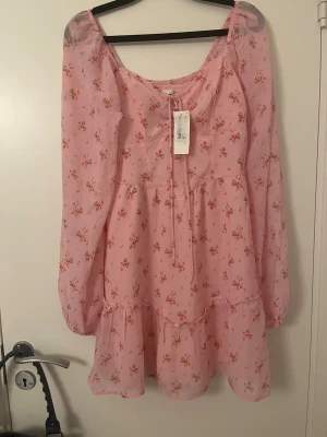 Rosa blommig kortklänning Hollister - Supersöt rosa kortklänning från Hollister med små blommor i rött och beige. Klänningen har långa, transparenta ärmar, snörning framtill och volangdetaljer vid kjolen. Smockad rygg för extra komfort och feminin passform. Perfekt för vår och sommar!