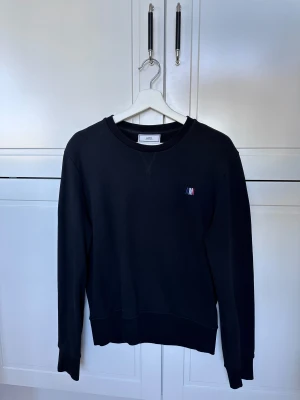 Svart sweatshirt från Ami Paris - Svart sweatshirt från Ami Paris med broderad logga i rött, vitt och blått på bröstet. Tröjan har rund hals, långa ärmar och ribbade muddar vid ärmslut och nederkant. Perfekt för en chill och stilren look.