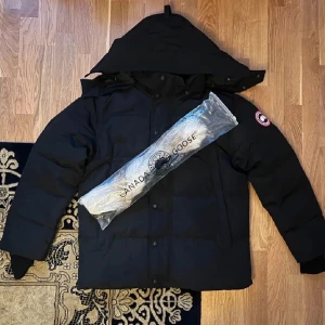 Svart dunjacka från Canada Goose - Säljer en svart dunjacka från Canada Goose med huva och klassisk logga på ärmen. Jackan har knappar framtill och är designad för att hålla dig varm under kalla dagar. Perfekt för dig som vill ha en stilren och funktionell vinterjacka. Pälsen kommer inte med därför sänker jag priser 