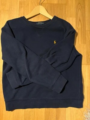 Mörkblå Ralph lauren tröja - Jag säljer en mörkblå polo Ralph lauren tröja med en snygg broderat märke i guld. Perfekt för en chill och stilren look. Snabb leverans.