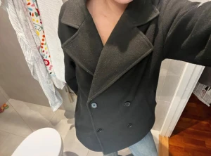 Svart skepparkavaj med bred krage - Säljer en svart skepparkavaj med bred krage och dubbelknäppning framtill. Jackan har en klassisk, rak passform och är tillverkad i ett ulligt material som känns mjukt och värmande