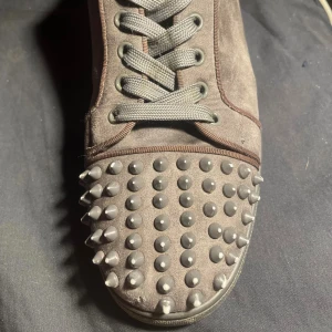 Christian Louboutin grå mocka sneakers - Säljer ett par gråa sneakers från Christian Louboutin i mocka med ikonisk röd sula och coola nitar på tån. Skorna har snörning och en platt sula, samt rund tå. Perfekt för dig som vill sticka ut med en edgy och lyxig stil.