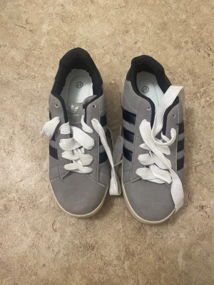 Adidas Campus grå sneakers strl 37 - Säljer ett par Adidas Campus sneakers i grå mocka med svarta ränder och vit sula. Skorna har vit snörning, svart häl och klassisk rund tå. Perfekt för dig som gillar retrostil och vill ha ett par snygga och bekväma sneakers.