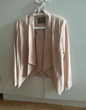 Puderrosa kavaj från Vero Moda - Säljer en snygg puderrosa kavaj från Vero Moda med öppen front utan knappar och snyggt draperad design. Kavajen har lång ärm och mjukt i polyester som ger ett stilrent och modernt intryck. Perfekt för att lyfta en outfit