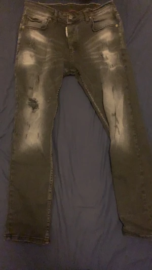 Svarta Dsquared2 jeans med slitningar W36 - Säljer ett par svarta jeans från Dsquared2 i storlek W36. Jeansen har coola slitningar och blekta detaljer framtill för en edgy look. Klassisk femficksmodell med knappgylf och Dsquared2-logga på insidan. Perfekta för dig som gillar streetstyle.