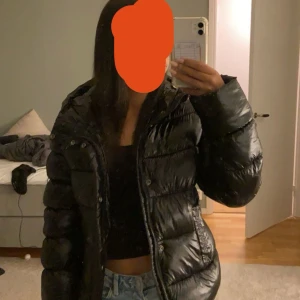 Svart pufferjacka med huva - Säljer en svart pufferjacka med glansig finish och huva. Jackan har långa ärmar, dragkedja och knappar framtill. Perfekt för kalla dagar och har en oversized look som är riktigt trendig just nu. Hål på som är lagat hos sömmerska 