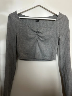 Grå croppad långärmad topp  - Säljer en grå croppad topp med långa ärmar och ribbad struktur. Toppen har en snygg rynkning framtill vid bysten och en rundad halsringning. Perfekt att styla med höga jeans eller kjol för en chill look.