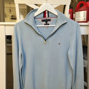 Ljusblå half zip tröja Tommy Hilfiger - Snygg ljusblå långärmad tröja från Tommy Hilfiger med half zip och klassisk krage med röd, vit och svart randig detalj. Tillverkad i mjuk bomull, perfekt för dig som gillar stilrena och sportiga vibes. Liten Hilfiger-logga broderad på bröstet.