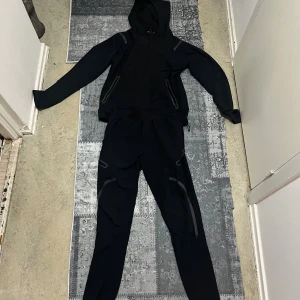 Svarta träningsbyxor Under Armour - Svarta träningsbyxor från Under Armour med gråa detaljer och dragkedjor på benen. Snyggt sportigt snitt, perfekt för dig som gillar att röra på dig. Materialet är syntet och byxorna har en slim passform med coola reflekterande inslag.