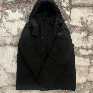 Wyndham black label jacka från Canada Goose  - Säljer en svart Canada Goose dunjacka med luva och avtagbar pälskant. Jackan har klassisk black label patch på ärmen, raka fickor och stängs med både dragkedja och knappar. Fodrad med dun och fjädrar för maxad värme. Perfekt för kalla dagar och riktigt snygg streetstil.