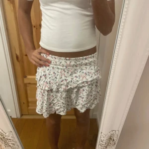 Vit blommig volangkjol från Cubus - Supersöt kort kjol i vit med små blommor i blått, rött och grått. Kjolen har volanger och ett smockat midjeband som gör den både bekväm och snygg. Perfekt för dig som gillar en feminin och lekfull stil.