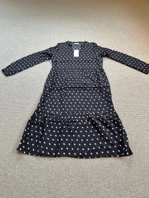 Svart prickig långklänning Noa Noa - Svart långklänning från Noa Noa med rosa och orange prickar, v-ringad hals och långa ärmar med resår vid ärmslut. Klänningen har volangdetalj nedtill och är tillverkad i mjuk viskos som faller snyggt. Perfekt för dig som gillar lekfulla mönster och sköna material.