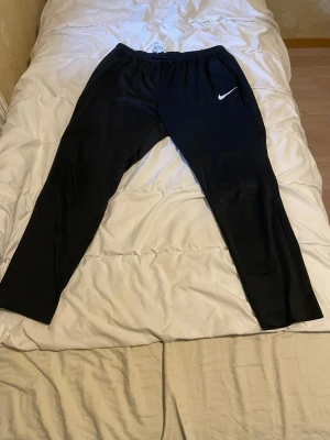 Svarta träningsbyxor från Nike - Svarta mjukisbyxor från Nike med klassisk vit logga på vänster lår. Byxorna har elastisk midja med snörning och är tillverkade i ett mjukt syntetmaterial som passar perfekt för träning. Passformen är normal och benen är raka.