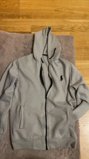 Grå hoodie från Polo Ralph Lauren - Snygg grå hoodie från Polo Ralph Lauren med svart broderad logga på bröstet. Tröjan har huva med snörning, hel dragkedja och två fickor framtill. Mjuk insida i bomullsmix och ribbade muddar vid ärmslut och nederkant. Perfekt för chill dagar.