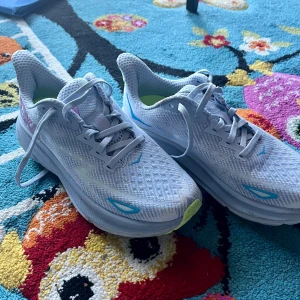 Hoka Clifton 9 joggingskor  - Säljer ett par helst nya Hoka Clifton 9 sneakers i ljusblått med detaljer i blått, lime och rosa. Skorna har mesh-ovandel för bra ventilation, snörning och uppbyggd sula. Perfekta för löpning eller träning, med bekväm dämpning och sportig look.