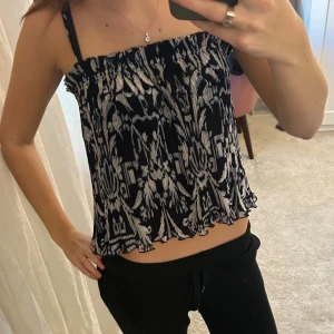Svartvit mönstrad linnetopp - Säljer en svartvit linnetopp med ett unikt mönster och plisserad struktur. Toppen har smala axelband och en croppad passform med vågig kant nedtill. Perfekt för dig som gillar coola detaljer och lätt material.