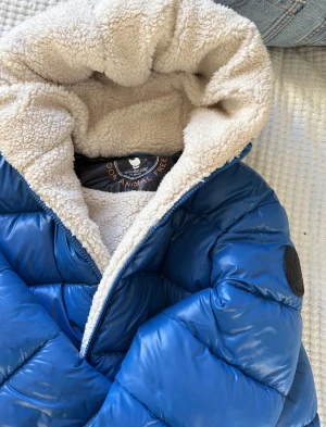 Blå pufferjacka med teddyfoder - Säljer en blå pufferjacka från Save the Duck med fluffigt teddyfoder i huvan och insidan. Jackan har dragkedja framtill, huva och en schysst loose passform. Perfekt för kalla dagar och riktigt mysig att bära. Fodret är animal free och jackan har en cool badge på ärmen.