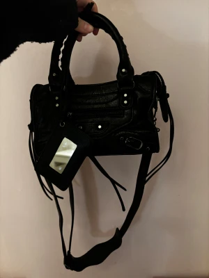 Svart liten handväska  - Söt svart handväska med silverfärgade detaljer och dragkedja. Väskan har justerbar axelrem, en spegel med inbyggd korthållare, och flera fack inuti. Perfekt storlek för det viktigaste och har snygga snördetaljer.