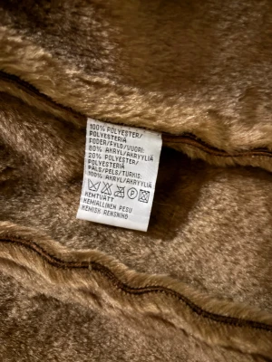 Brun pälsjacka från Fashion Quality - Säljer en brun pälsjacka med fluffiga detaljer i beige och ljusbrunt längs krage, ärmslut och nederkant. Jackan har långärmad design och är gjord i polyester och akryl för en mjuk och varm känsla. Perfekt för dig som vill ha en cool retrostil med statementkrage.