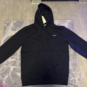 Svart hoodie från Comme des Garçons Play - Svart hoodie från Comme des Garçons Play i storlek S. Tröjan har huva med snörning, en klassisk känguruficka och det ikoniska hjärteögon-logotypen på bröstet. Mjuk bomullskvalitet och långärmad modell, perfekt för en chill och stilren look.