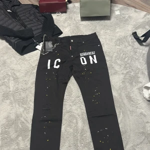 Svarta Dsquared2 ICON Black Bull jeans - Säljer ett par svarta Dsquared2 ICON Black Bull jeans med vit text och coola färgstänk i gult och vitt på benen. Jeansen har klassisk femficksdesign, silverfärgade knappar och detaljer, samt Dsquared2-märke i midjan. Tillverkade i Italien.