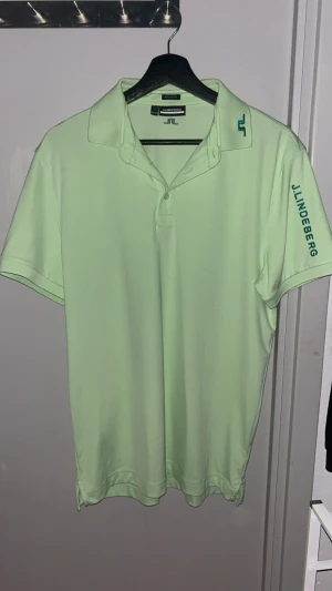 Ljusgrön J.Lindeberg piké - Fräsch ljusgrön pikétröja från J.Lindeberg med korta ärmar och klassisk krage. Diskret logga på kragen och ryggen samt J.Lindeberg-tryck på ena ärmen. Skön och lätt polyester som passar perfekt när du vill vara både chill och sportig.