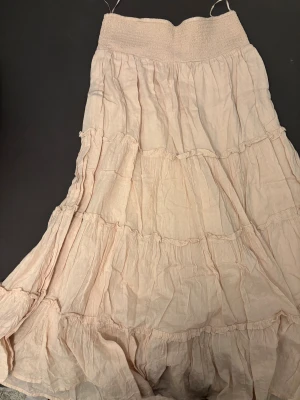 Beige volangkjol i bomull - Lång, ljusbeige maxikjol med flera volanger och smock i midjan. Kjolen är tillverkad i tunn bomull med diskret strukturmönster och har en luftig, flowy känsla. Perfekt för dig som gillar en bohemisk och avslappnad stil.