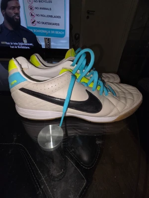 Nike Tiempo vit/blå fotbollsskor stl 41 - Nike Tiempo fotbollsskor i vitt/beige med turkos swoosh-loggor, blå skosnören och detaljer i neon-gult. Skorna har quiltad ovandel i skinn, platt gummisula med gul mönstrad undersida och rund tå. Perfekt för inomhusspel i stl 41 använda fåtal gånger.