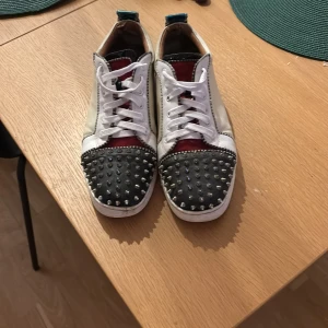 Christian Louboutin sneakers med nitar(röd undersida) - Unika sneakers från Christian Louboutin med vit skinnbas, svart tåparti täckt av silvriga nitar, röda detaljer och metallicblå häl. Röd undersida, snygg snörning och klassisk röd sula. Perfekt för dig som vill sticka ut med en edgy och lyxig stil. Nypris 7500-8000kr  Skriv privat för mer information! 