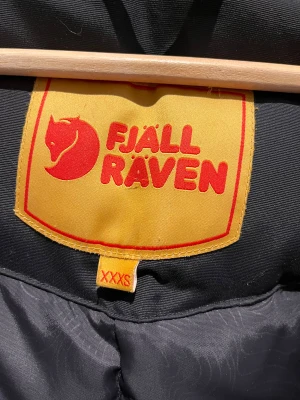 Svart dunjacka från Fjällräven XXXS - Svart dunjacka från Fjällräven med klassisk röd logga på bröstet och gul insida vid etiketten. Jackan har huva, flera fickor med lock och dragkedja framtill. Perfekt för kalla dagar och har en schysst, puffig look som är både praktisk och snygg.  XXXS herr storlek motsvarar storleken S