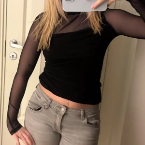 Svart mesh topp med långa ärmar - Säljer en svart mesh topp med långa ärmar och transparenta partier över axlar och armar. Toppen är figurnära och har en hög hals. Perfekt för dig som gillar en edgy och trendig stil.