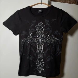 Svart t-shirt med kors från Urban Outfitters Affliction - Svart t-shirt från Urban Outfitters med ett stort, detaljerat kors i silver på framsidan. T-shirten har rund hals, korta ärmar och är gjord i mjuk bomull. Snyggt tryck med ornament och silvriga detaljer som ger en edgy vibe.