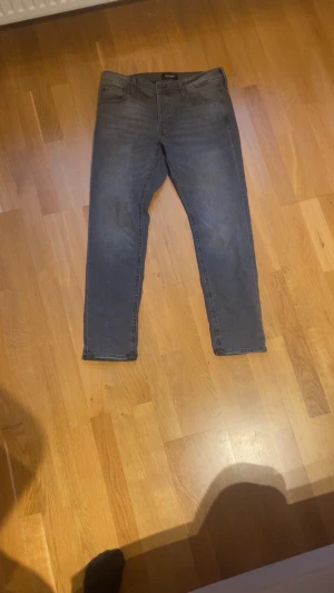 Grå jeans från Jack & Jones - Snygga grå jeans från Jack & Jones, modell Slim Glenn. De har en slim passform med klassisk femficksdesign och svarta detaljer vid knapparna. Jeansen är tillverkade i stretchigt denim för extra komfort och har en mörkgrå tvättad look.