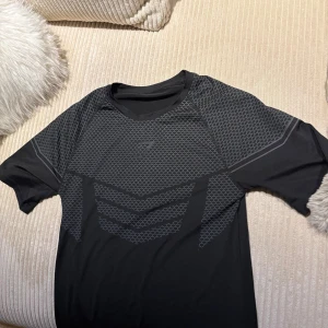 Svart Gymshark onyx v2 - Snygg svart t-shirt från Gymshark med grått geometriskt mönster över bröstet och axlarna. T-shirten har korta ärmar och är gjord i ett lätt och stretchigt polyestermaterial som andas, perfekt för träning. Diskret Gymshark-logga framtill.
