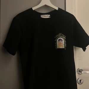 Svart t-shirt Casablanca S - Svart t-shirt från Casablanca Tennis Club i storlek S. Tryck fram och bak med motiv av en byggnad i fönsterdesign, omgiven av gröna blad och texten 'Avenida Casablanca' i gult. Klassisk rund hals och korta ärmar. Materialet är mjuk bomull.
