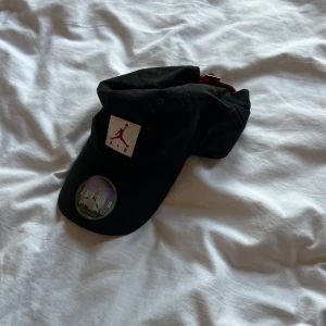 Svart Jordan keps med röd logga - Svart keps från Jordan med klassisk Jumpman-logga i rött framtill. Kepsen har böjd skärm, justerbar rem baktill med röd insida och är tillverkad i bomull. Perfekt för dig som vill ha en sportig och stilren look.