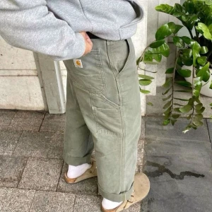 Gröna cargopants från Carhartt - Snygga gröna cargopants från Carhartt med bred passform och flera praktiska fickor. Byxorna har robusta sömmar, hammarögla på sidan och klassisk Carhartt-logga på bakfickan. Tillverkade i slitstark bomull, perfekta för en chill streetwear-look. Första bilden är inte min
